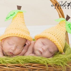 May include: Two newborn babies wearing yellow knitted pear hats with green leaves and the words "Perfect" and "Pair" on them. The babies are sleeping in a wicker basket lined with green fur.