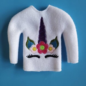 May include: White fleece elf shirt with an embroidered unicorn design. The unicorn has a purple horn, green leaves, a pink flower, and yellow flowers.