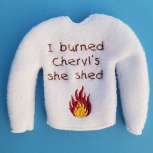 May include: White elf-sized sweater with the text "I burned Cheryl's she shed" and a red and yellow embroidered flame.