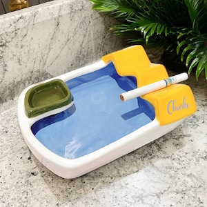May include: A ceramic ashtray in white, blue, and yellow. The rectangular ashtray features a blue interior, a small green dish, and the name "Charlie" in blue script. A lit cigarette rests on the side of the ashtray.
