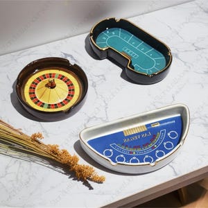 Könnte beinhalten: Drei Aschenbecher aus Keramik mit Casino-Motiven. Ein Aschenbecher zeigt ein Roulette-Rad, ein anderer einen grünen Filz-Pokertisch und der dritte einen blauen Filz-Blackjack-Tisch mit dem aufgedruckten Text "LAS VEGAS".