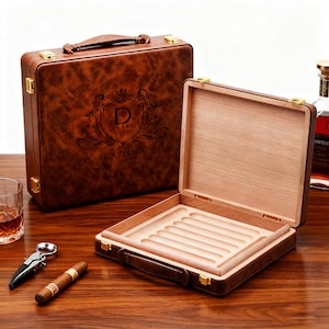 May include: A brown leather cigar humidor with gold-colored hardware, open to reveal a wooden interior with cigar slots. The closed humidor features a decorative crest with the text "D TAYLOR 1978". A cigar, cutter, and glass of whiskey are also visible.
