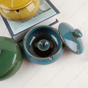 Könnte beinhalten: Drei Aschenbecher aus Keramik, einer gelb, einer grün und einer blau mit Deckel. Der blaue Aschenbecher hat ein dunkelblaues Inneres.