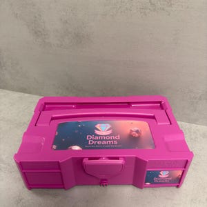 May include: A vibrant pink storage box featuring the text "Diamond Dreams" and a diamond emblem. The box includes a handle and a latch. It is designed for storing diamond art supplies, with the slogan "Store the Bling, Create the Dream."
