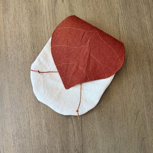 May include: A small fabric pouch featuring a cream-colored base and a rust-colored flap shaped like a leaf. The leaf-shaped flap includes stitched vein details and is secured with a thin brown cord that ties at the front of the pouch.