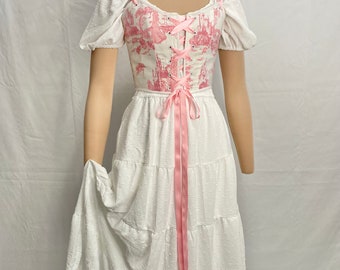 May include: A white tiered skirt with a pink ribbon tied at the waist. The skirt is paired with a white and pink corset top with a lace-up front and a toile pattern.