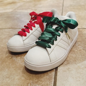 May include: A pair of white sneakers with green and red satin ribbon laces. The laces are tied in a bow on each shoe.