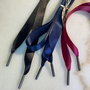 May include: Close-up of four pairs of shoelaces in different colors: black, navy blue, and burgundy. Each pair has a metal aglet at the end. The navy blue laces are tied together with twine.