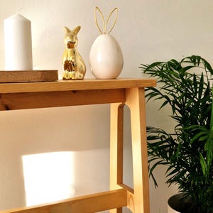 Pode incluir: Uma mesa de madeira com uma vela branca, uma figura de coelho dourado e um vaso branco em forma de ovo com orelhas de coelho douradas. Uma planta em vaso está à direita. Os objetos criam um tema de primavera ou Páscoa.