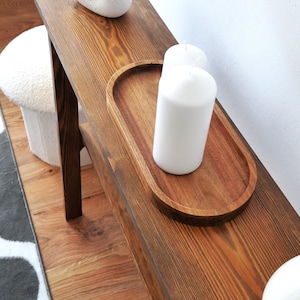 Puede incluir: Una mesa consola de madera con un tinte marr&oacute;n oscuro. La mesa tiene una tapa rectangular y dos patas. Hay tres velas blancas en una bandeja de madera sobre la mesa.