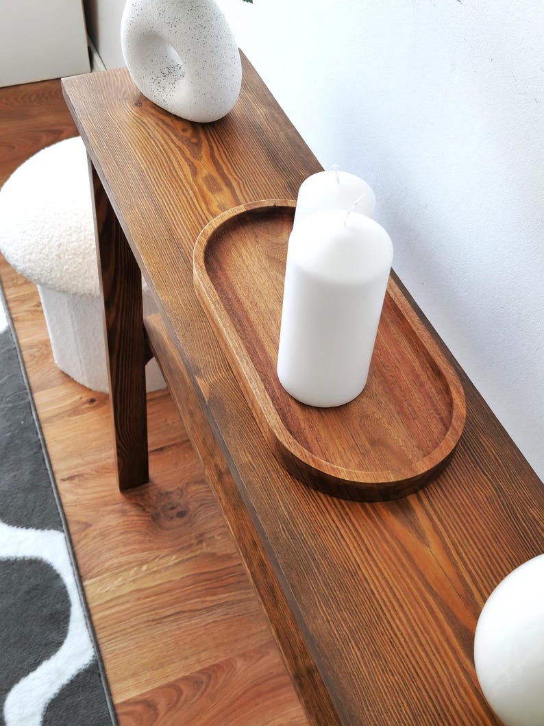 Puede incluir: Una mesa consola de madera con un tinte marr&oacute;n oscuro. La mesa tiene una tapa rectangular y dos patas. Hay tres velas blancas en una bandeja de madera sobre la mesa.