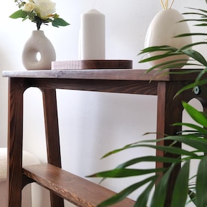 Puede incluir: Una mesa consola de madera marr&oacute;n oscuro con dos estantes. La mesa tiene un jarr&oacute;n de cer&aacute;mica blanca con flores blancas, una vela blanca y un conejito de cer&aacute;mica blanca con orejas doradas en la parte superior.
