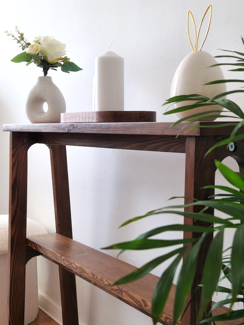 Puede incluir: Una mesa consola de madera marr&oacute;n oscuro con dos estantes. La mesa tiene un jarr&oacute;n de cer&aacute;mica blanca con flores blancas, una vela blanca y un conejito de cer&aacute;mica blanca con orejas doradas en la parte superior.