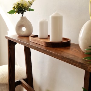 Może przedstawiać: Brązowy drewniany stolik konsolowy z białym ceramicznym wazonem z otworem z boku, wypełnionym białymi kwiatami. Dwie białe świece znajdują się na drewnianej tacy na stole.