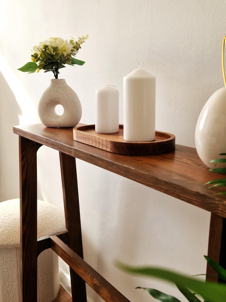Puede incluir: Una mesa consola de madera marr&oacute;n oscuro con un jarr&oacute;n de cer&aacute;mica blanca con un agujero en el lateral, lleno de flores blancas. Dos velas blancas est&aacute;n en una bandeja de madera sobre la mesa.