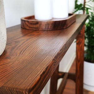 Puede incluir: Una mesa consola de madera marr&oacute;n oscuro con un acabado de veta de madera natural. La mesa tiene una tapa rectangular y un dise&ntilde;o simple y moderno. La mesa est&aacute; decorada con una bandeja de madera y dos velas blancas.