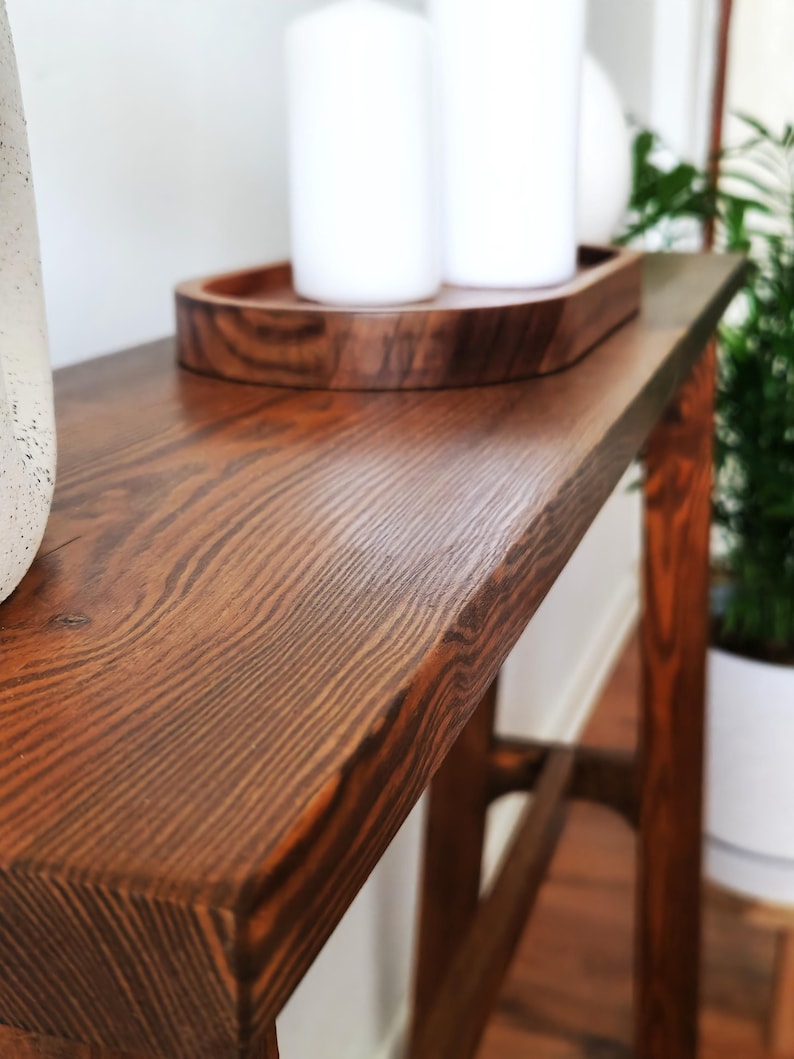 Puede incluir: Una mesa consola de madera marr&oacute;n oscuro con un acabado de veta de madera natural. La mesa tiene una tapa rectangular y un dise&ntilde;o simple y moderno. La mesa est&aacute; decorada con una bandeja de madera y dos velas blancas.