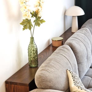 Puede incluir: Interior de salón con una estantería de madera oscura que exhibe un jarrón de vidrio verde texturizado con flores color crema. Una lámpara con forma de hongo y una vela están sobre la estantería. En primer plano, un sofá de pana gris con un cojín con estampado de leopardo.