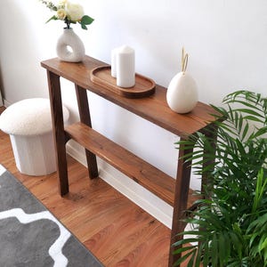 Puede incluir: Una mesa consola de madera marr&oacute;n oscuro con dos estantes. La mesa tiene un jarr&oacute;n de cer&aacute;mica blanca con flores blancas, dos velas blancas y un objeto decorativo de cer&aacute;mica blanca en la parte superior. La mesa est&aacute; frente a una pared blanca y una alfombra de patr&oacute;n gris y blanco.