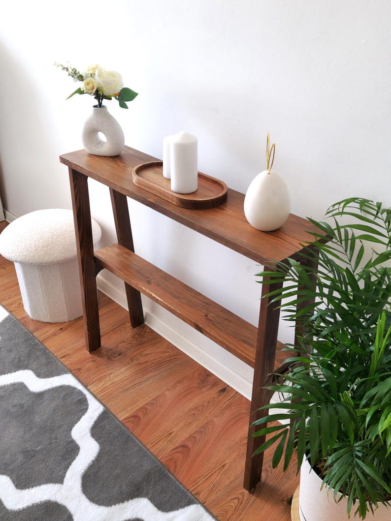 Puede incluir: Una mesa consola de madera marr&oacute;n oscuro con dos estantes. La mesa tiene un jarr&oacute;n de cer&aacute;mica blanca con flores blancas, dos velas blancas y un objeto decorativo de cer&aacute;mica blanca en la parte superior. La mesa est&aacute; frente a una pared blanca y una alfombra de patr&oacute;n gris y blanco.