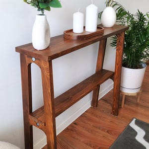 Puede incluir: Una mesa consola de madera marr&oacute;n oscuro con un estante. La mesa tiene un jarr&oacute;n blanco con flores blancas, tres velas blancas en una bandeja de madera y un huevo de cer&aacute;mica blanco con detalles dorados en la parte superior.