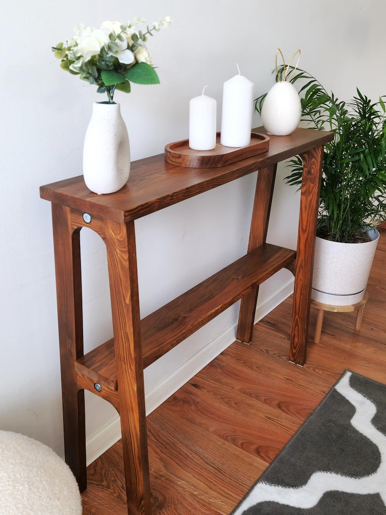Puede incluir: Una mesa consola de madera marr&oacute;n oscuro con un estante. La mesa tiene un jarr&oacute;n blanco con flores blancas, tres velas blancas en una bandeja de madera y un huevo de cer&aacute;mica blanco con detalles dorados en la parte superior.