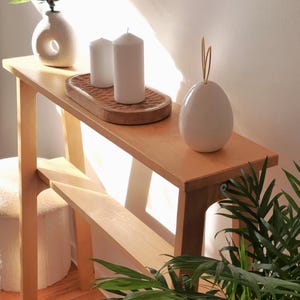 Puede incluir: Una consola de madera clara con un jarrón de cerámica blanca con flores blancas, dos velas blancas, una bandeja de madera y un objeto de cerámica blanca en forma de huevo con un tallo de alambre dorado. La mesa tiene un estante inferior y está en una habitación con un suelo de madera clara y una planta verde.