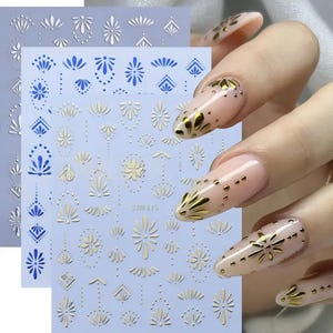 May include: Nail art stickers in gold and blue, featuring floral and geometric designs. The stickers are on a light blue background. The image also shows nails with gold nail art.
