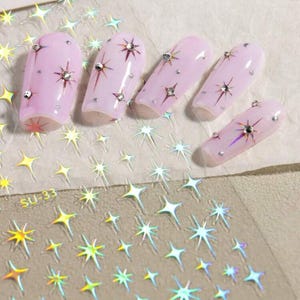 May include: A sheet of iridescent nail stickers with a design of stars. The sheet is labeled "SU-33". The stickers are shown applied to pink nail tips.