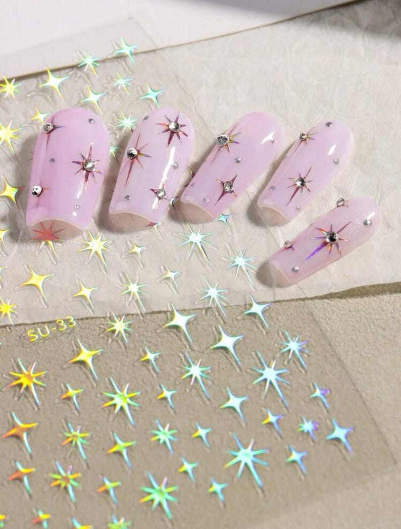 May include: A sheet of iridescent nail stickers with a design of stars. The sheet is labeled "SU-33". The stickers are shown applied to pink nail tips.