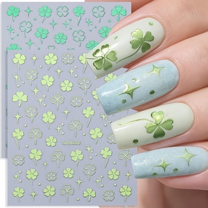 May include: Nail art stickers with green shamrocks and stars on a grey background. The stickers are applied to nails with a light green shamrock design and light blue nails with star designs. The text "SWA200" is visible on the sticker sheet.
