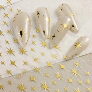May include: Gold nail art stickers with a starburst design. The stickers are on a clear sheet with a white background. The sheet has the text "SU-233" printed on it.