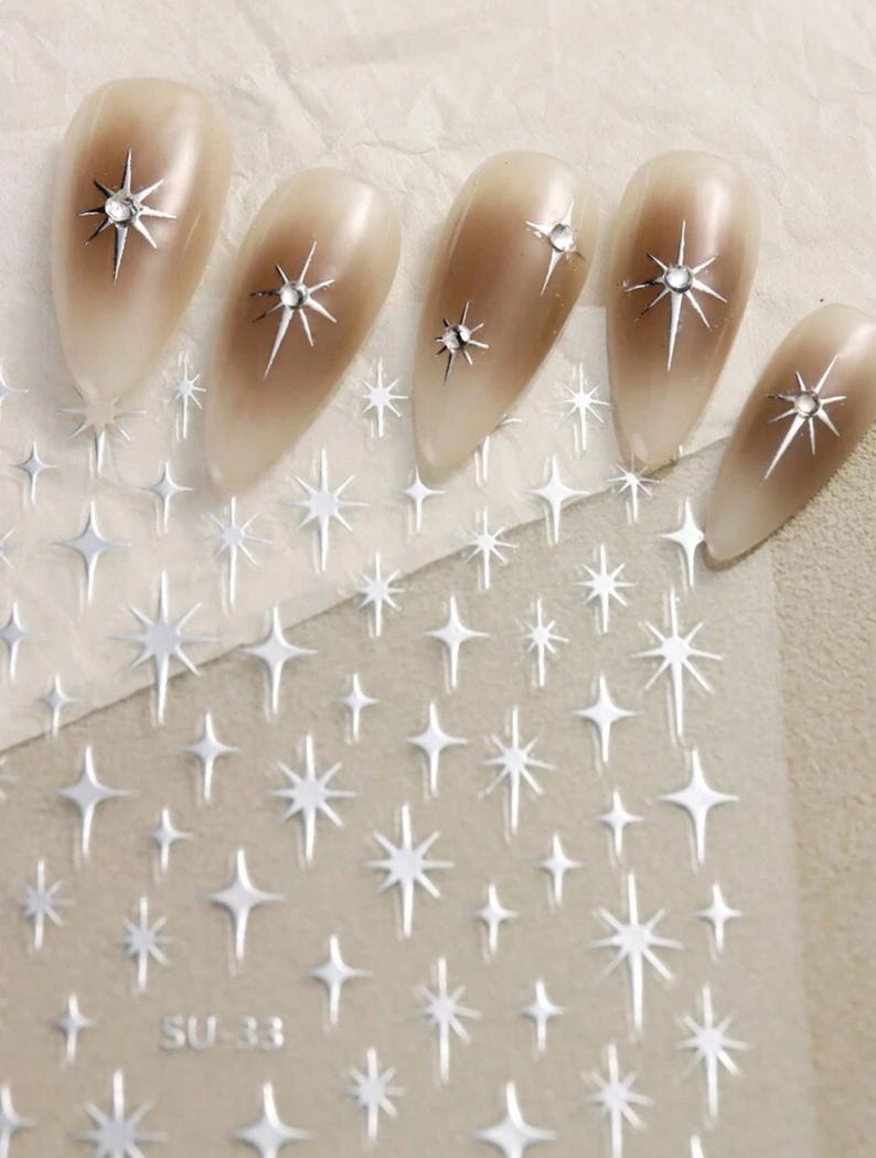 May include: Nail art stickers with a silver glitter star design. The sheet includes multiple stars of varying sizes. The sheet also has the text "SU-33" printed on it.