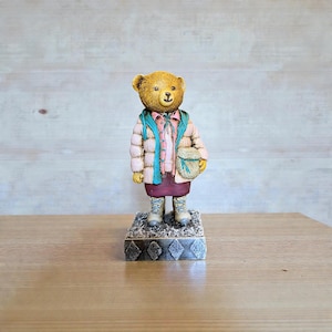 Könnte beinhalten: Dekorative Bärenfigur mit einer rosa Steppjacke, einem türkisfarbenen Schal und einem weinroten Rock. Der Bär hält eine kleine Tasche und steht auf einem dekorativen Sockel. Die Figur hat ein detailliertes Design und besteht aus einem haltbaren Material.