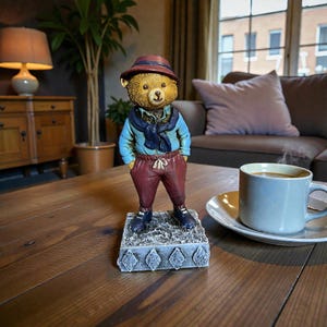 Könnte beinhalten: Dekorative Bärenfigur mit Hut, blauem Hemd, weinroter Hose und blauem Schal. Der Bär steht auf einem dekorativen Sockel. Die Figur hat eine Vintage-Ästhetik und steht auf einem Holztisch.