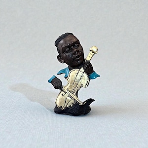 May include: A figurine of a Black musician playing a violin. The figure is wearing a blue shirt and has a gold-colored violin. The base is black with gold accents. The figurine is on a light gray surface.