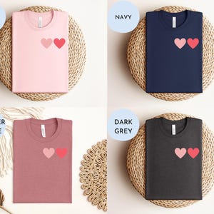 May include: Four t-shirts with a two-heart graphic design. The shirts are shown in pink, navy, heather mauve, and dark grey.