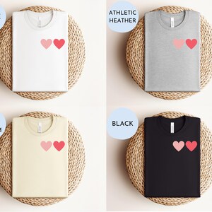 May include: Four t-shirts with a graphic of two pink hearts on the left chest. The t-shirts are displayed on a woven background. The t-shirt colors are white, athletic heather, soft cream, and black.