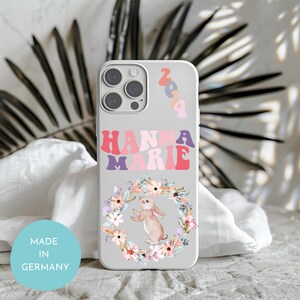 May include: A white phone case featuring a pink, purple and blue floral wreath with a bunny in the centre. The text "HANNA MARIE" is in a retro font above the wreath. The year "2014" is in the top right corner.