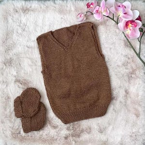 Peut inclure: Un gilet marron tricoté avec un col en V et des chaussons assortis. Le gilet est de couleur marron moyen et semble fait à la main. Les chaussons sont petits et également marron. Les articles sont présentés sur une surface blanche et moelleuse.