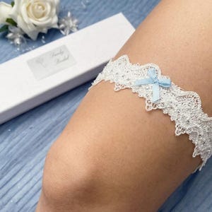 May include: A white lace garter featuring a small light blue ribbon bow at the center. The lace has a scalloped edge and is embellished with tiny clear beads. It is presented with a white rectangular gift box labeled Truly Bridal.