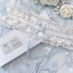 May include: A white lace bridal garter with floral details and a light blue ribbon. The garter is adorned with faux pearls and rests on a white box with the text "Purely Bridal". Decorative white roses and pearls are in the background.