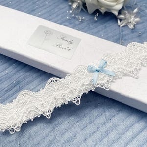May include: A white lace garter featuring a small light blue ribbon bow, resting on a white rectangular gift box. The box displays a label with a rose illustration and the text Ferdy Bodel. The lace has a scalloped edge and intricate detailing.