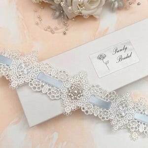 May include: A white lace wedding garter with light blue satin ribbons and a central pearl and rhinestone embellishment. The garter is displayed on a white box with the text "Purely Bridal".