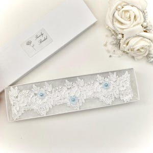 May include: A white lace garter with light blue floral details, presented in a clear box. The garter showcases detailed leaf and floral designs. A white box with the text "Purely Bridal" is also visible.