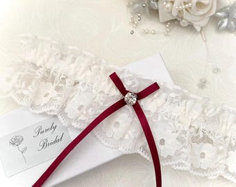 May include: White lace garter with a burgundy satin bow and a rhinestone embellishment. The garter is on a white box with the text "Purely Bridal".