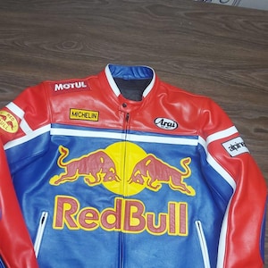 May include: Red, blue, and white leather jacket with the Red Bull logo. The jacket has a zipper closure and multiple sponsor logos, including Michelin and Arai. The jacket is a racing style with a stand-up collar.