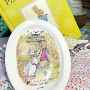 May include: A white ceramic dish with an oval shape, featuring a colorful illustration from "The Tale of Jemima Puddle-Duck" by Beatrix Potter. A yellow book titled "The Tale of Peter Rabbit" is in the background.