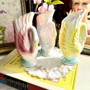 May include: Three iridescent ceramic swan vases with a pastel pink, blue, and yellow glaze. The vases are sitting on a book with a yellow cover and the text "Alice's Adventures in Wonderland".