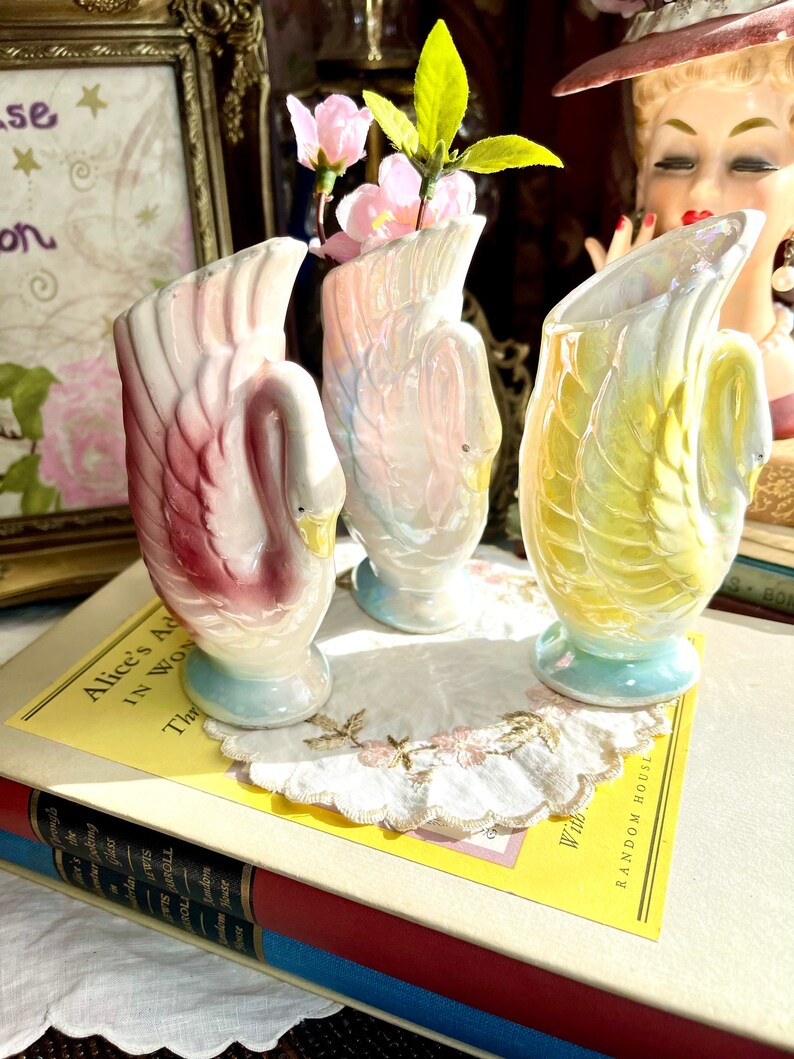 May include: Three iridescent ceramic swan vases with a pastel pink, blue, and yellow glaze. The vases are sitting on a book with a yellow cover and the text "Alice's Adventures in Wonderland".